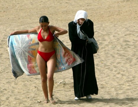 A young woman wearing a bikini walks along the beach with a friend in dressed in traditional clothes at the Palm Beach Club in Algiers July 25, 2003. [It is the summer of 2003 and the scars from a decade of Islamist extremist-linked violence that left between 100,000 and 150,000 people dead in the oil-rich north African country are almost no longer visible.] Temperatures soar above 40 degrees Celsius and people are back on Algeria's beaches escaping the town's heat and having fun